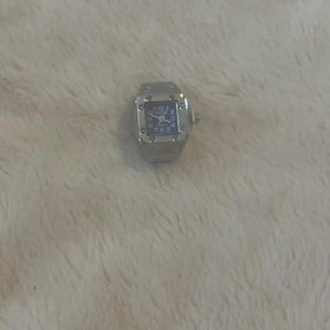 May include: A silver watch ring with a square face and a blue dial. The watch has a metal band and a small crown. The watch face displays the time with silver hands and numerals. The ring is set against a white, textured background.