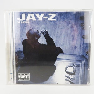 Puede incluir: Una caja de CD con un fondo azul y blanco que presenta a un hombre sentado en una superficie de madera. El texto "JAY-Z THE BLUEPRINT" está impreso en la parte frontal de la caja. Una pegatina de advertencia para padres está en la esquina inferior derecha.