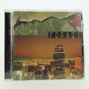 Op de afbeelding: Een CD-hoes met een zwarte rug en een voorkant met een nachtelijke stadsgezicht met een hoog gebouw in het midden. De tekst "FUGAZI" is in gouden letters op de hoes gedrukt.