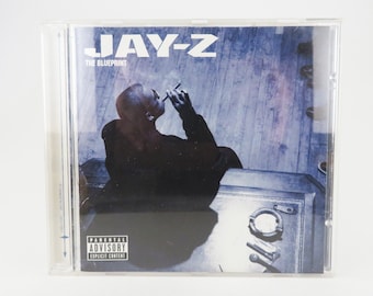May include: A CD case with a blue and white background featuring a man sitting on a wooden surface. The text "JAY-Z THE BLUEPRINT" is printed on the front of the case. A parental advisory sticker is in the bottom right corner.