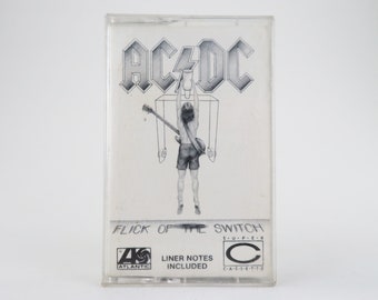 May include: A cassette tape case with a black and white image of a man holding a guitar. The text "AC/DC" is printed in large, bold letters at the top of the case. The text "FLICK OF THE SWITCH" is printed below the image. The text "SUPER" is printed in smaller letters below the album title. The text "LINER NOTES INCLUDED" is printed below the album title. The text "ATLANTIC" is printed in a circle below the album title. The text "C-A-S-S-E-T-T-E" is printed in a circle below the album title.