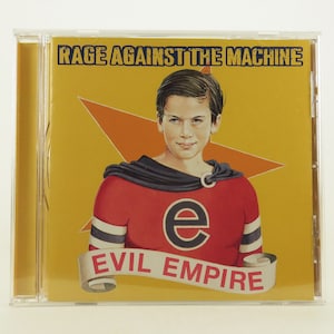 Puede incluir: Una caja de CD con fondo amarillo y una estrella roja. La caja presenta un personaje de superhéroe con un traje rojo y negro y el texto "Evil Empire" en una pancarta. El texto "Rage Against The Machine" está impreso en negro en la parte superior de la caja.
