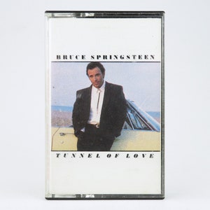 Puede incluir: Una cinta de casete con una imagen en blanco y negro de Bruce Springsteen de pie frente a un coche amarillo. El texto "Bruce Springsteen" y "Tunnel of Love" está impreso en la cinta de casete.