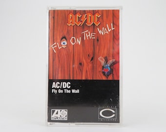 May include: A cassette tape with a red, white, and black cover. The cover features a wooden wall with a fly and an eye painted on it. The text "AC/DC Fly On The Wall" is printed on the cover.