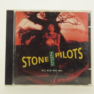 May include: A red and black CD case with a photo of a person standing in front of a tree. The text "Stone Temple Pilots" and "Core" are printed on the case.