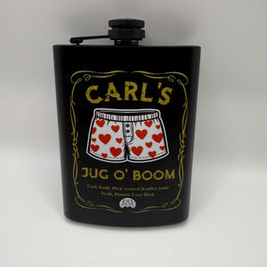 Peut inclure: Une flasque en métal noir avec un bouchon à vis. L'avant présente une bordure décorative jaune, le texte "CARL'S JUG O' BOOM", une illustration de caleçon blanc à cœurs rouges et le texte "Carl, look, they named it after you! Yeah, Donut. I see that."