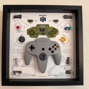 May include: A black shadow box frame containing a disassembled grey Nintendo 64 controller. The display features the controller shell, internal circuit boards, buttons, and joysticks arranged on a white background with technical labels and diagrams.