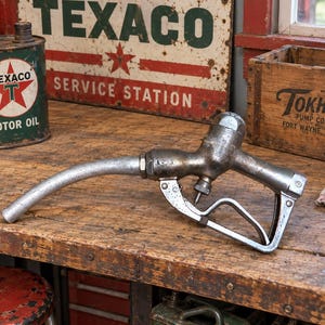 May include: A vintage metal fuel nozzle with a curved spout, a trigger mechanism, and a handle guard. The industrial tool features a weathered, dark gray finish with visible signs of age, oxidation, and metallic texture throughout its structure.