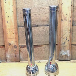 May include: Two vintage chrome flashlights with ribbed bases and red power buttons. The flashlights are cylindrical with a slightly tapered design. They are set against a wooden background.