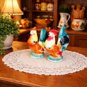 May include: Two ceramic rooster figurines with vibrant colors of yellow, orange, red, and blue. The roosters are perched on a light blue base, set on a white lace doily. The background includes a wooden cabinet and various decorative items.