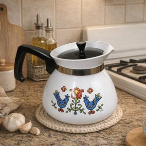 May include: White enamel coffee pot with a black handle and lid, featuring a gold band and a colorful floral and bird design. The pot sits on a beige crocheted coaster, with a stove and kitchen utensils in the background.