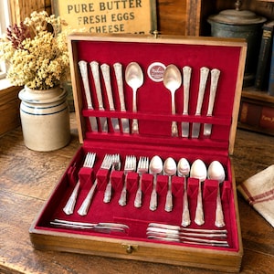 May include: A wooden flatware storage chest with a deep red velvet interior lining, containing a complete set of silver-toned metal utensils including various spoons, forks, and knives arranged in dedicated slots, with a small circular label.