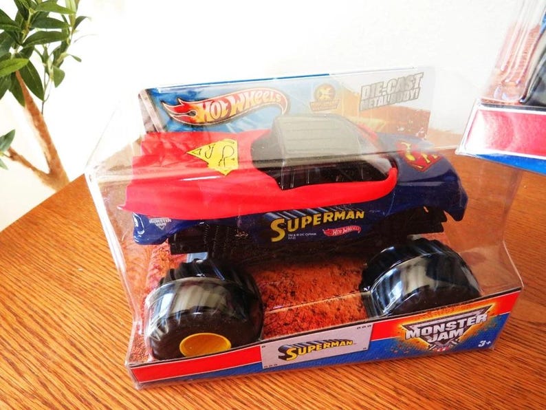 May include: A Hot Wheels Superman monster truck toy in a clear plastic package. The truck is blue with a red hood and the Superman logo. The packaging includes the words "Superman", "Hot Wheels", and "Monster Jam".
