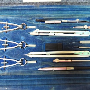 May include: A vintage drafting set featuring various metal compasses, dividers, and technical drawing instruments arranged in a blue velvet-lined case. The collection includes multiple silver-toned tools with adjustable screws and black handles.
