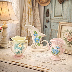 May include: Three vintage ceramic vases in pastel colors. The vases feature floral designs with blue flowers and green leaves. One vase is light pink, another is cream, and the third is pale yellow. The vases are displayed on a lace tablecloth.