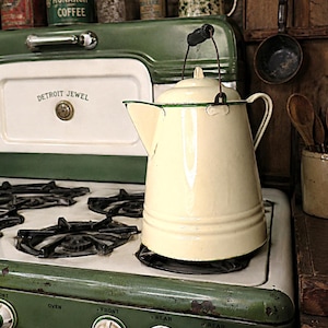 May include: A cream-colored enamel coffee pot with a green rim and a black handle sits on a vintage green and white Detroit Jewel stove. The stove features multiple black metal burners, control knobs, and a white panel with the text Detroit Jewel.