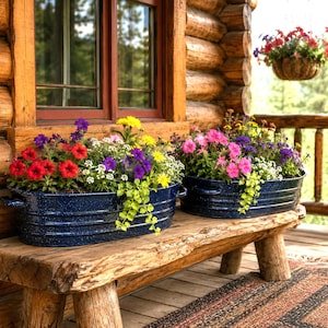 May include: Two oval, navy blue speckled enamel metal tubs with side handles, each filled with a vibrant arrangement of red, purple, yellow, and white flowers, resting on a rustic wooden bench in front of a log cabin window.