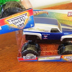 May include: A blue and white die-cast metal Grave Digger monster truck toy in its original packaging. The packaging features the Monster Jam logo and the text "Grave Digger Legend". The toy is suitable for ages 3+.
