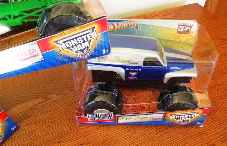 May include: A blue and white die-cast metal Grave Digger monster truck toy in its original packaging. The packaging features the Monster Jam logo and the text "Grave Digger Legend". The toy is suitable for ages 3+.