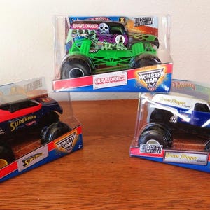 May include: Three die-cast Monster Jam trucks in their original packaging. The trucks are a green Grave Digger, a blue and red Superman truck, and a white and blue Grave Digger truck. The packaging features the Monster Jam logo.
