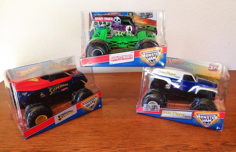 May include: Three die-cast Monster Jam trucks in their original packaging. The trucks are a green Grave Digger, a blue and red Superman truck, and a white and blue Grave Digger truck. The packaging features the Monster Jam logo.