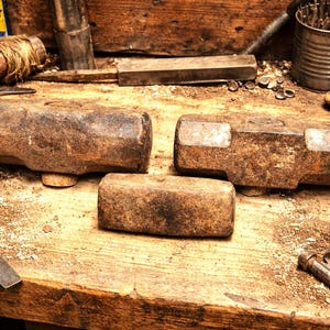 May include: A collection of well-worn blacksmithing tools, including three large, rectangular sledgehammers with wooden handles, arranged on a wooden workbench. Other tools include a chisel, metal fasteners, and a tin can filled with metal rods.