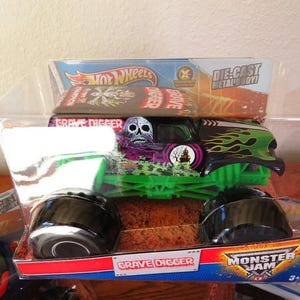 May include: A Hot Wheels Grave Digger monster truck toy in its original packaging. The die-cast metal body truck features a skull graphic, green flames, and a green chassis. The packaging includes the Monster Jam logo.