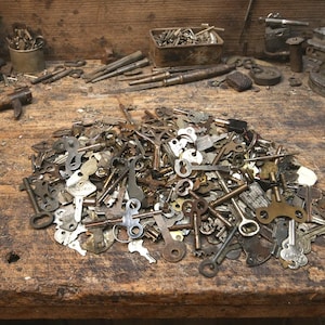 May include: A large pile of antique keys of various shapes and sizes rests on a weathered wooden workbench. The keys are made of metal and show signs of age and wear. Other tools and hardware are scattered around the workbench.