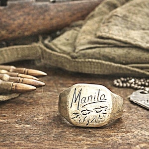 May include: A vintage ring with the word "Manila" engraved on it, along with the year "1944". The ring is placed on a wooden surface, alongside bullets, a military tag, and a canvas bag.