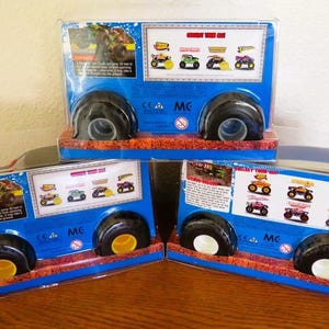 May include: Three toy monster truck packages. Each package is blue with black wheels and a clear plastic front. The packages have images of different monster trucks and text. The wheels have yellow and white accents.