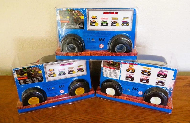 May include: Three toy monster truck packages. Each package is blue with black wheels and a clear plastic front. The packages have images of different monster trucks and text. The wheels have yellow and white accents.