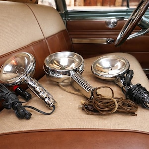 May include: Three vintage chrome spotlights with textured handles and coiled cords rest on a tan and brown car seat. The spotlights have a reflective bowl shape, suggesting they are for automotive use.