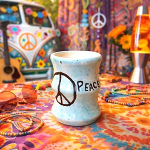 May include: A light blue ceramic mug with the word "Peace" and a peace symbol. The mug is on a colorful patterned surface with beaded necklaces and orange sunglasses. A vintage van and guitar are in the background.