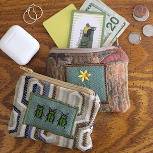 Puede incluir: Dos pequeños monederos de tela estampada con cierre de cremallera. Un monedero tiene un rectángulo verde bordado con tres escarabajos verdes. El otro tiene un bordado de flor amarilla. Se ve dinero y monedas.