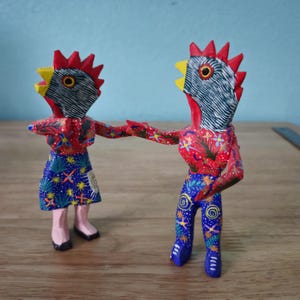 Puede incluir: Dos coloridas figuras de gallos de madera pintadas a mano. Una lleva una falda azul con diseños de estrellas y flores, la otra lleva pantalones azules con patrones similares. Ambos tienen crestas rojas, picos amarillos y se dan la mano.