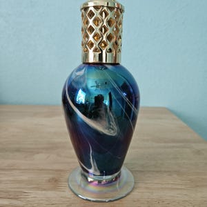 Puede incluir: Una lámpara de fragancia con cuerpo de vidrio en forma de pera que presenta un acabado iridiscente azul y morado con vetas blancas. La parte superior está cubierta con una tapa de metal dorado con un patrón de corte en forma de diamante.