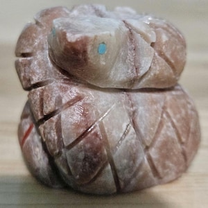 May include: A carved stone sculpture of a snake with turquoise eyes. The sculpture is a light brown colour with carved details and a textured surface. The snake is coiled and appears to be made of a single piece of stone.