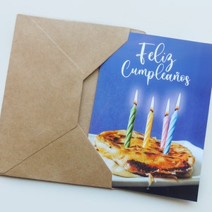 May include: A birthday card with a blue background and the text "Feliz Cumplea&ntilde;os". The card features a brown pastry with four lit candles, two green, one pink, and one blue.