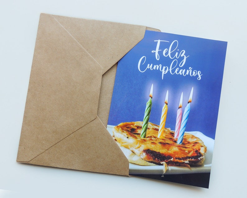 May include: A birthday card with a blue background and the text "Feliz Cumplea&ntilde;os". The card features a brown pastry with four lit candles, two green, one pink, and one blue.
