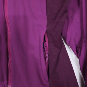May include: A close-up of a purple jacket with a full zipper and a zippered pocket. The jacket has a textured fabric and a darker purple panel on the side. The zipper and pocket are a lighter shade of purple.