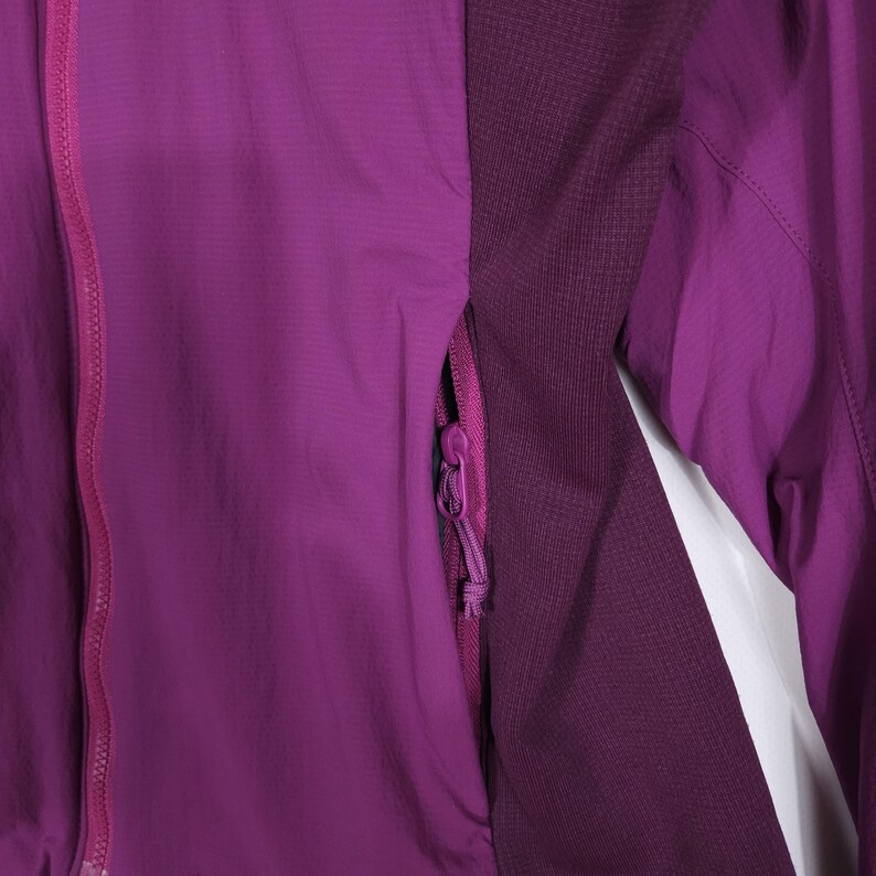 May include: A close-up of a purple jacket with a full zipper and a zippered pocket. The jacket has a textured fabric and a darker purple panel on the side. The zipper and pocket are a lighter shade of purple.