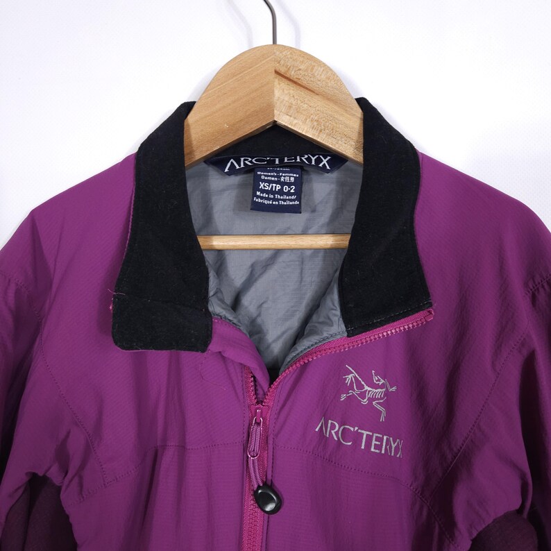 May include: A purple Arc'teryx jacket with a black collar and a full zipper. The jacket has the Arc'teryx logo on the front and is hanging on a wooden hanger. The jacket is a size XS/TP 0-2.