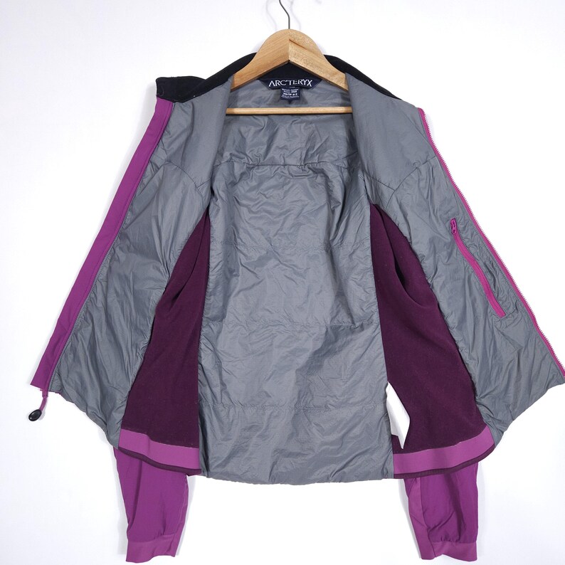 May include: Open view of a purple and black Arc'teryx jacket hanging on a wooden hanger. The jacket has a black collar, a gray interior lining, and a purple exterior with a zipper and pockets. The Arc'teryx logo is visible.