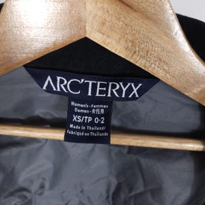 May include: Close-up of a black garment tag with the brand name ARC'TERYX in white. The tag includes text in English, German, and Japanese, along with the size XS/TP 0-2. The tag is attached to a black garment.