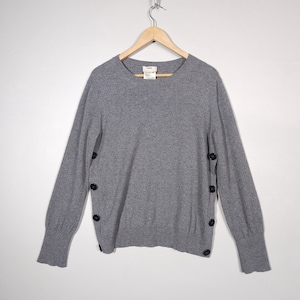 May include: A gray long-sleeved sweater with black buttons down the sides of the sleeves. The sweater has a round neckline.
