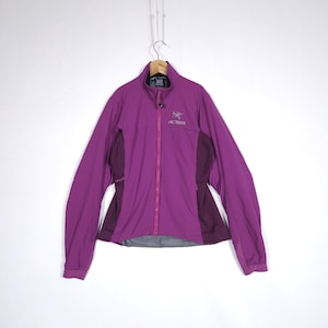 May include: A purple jacket with a full-length zipper and the brand name "Arc'teryx" on the chest. The jacket has a high collar and dark purple panels on the sides. It is hanging on a wooden hanger.