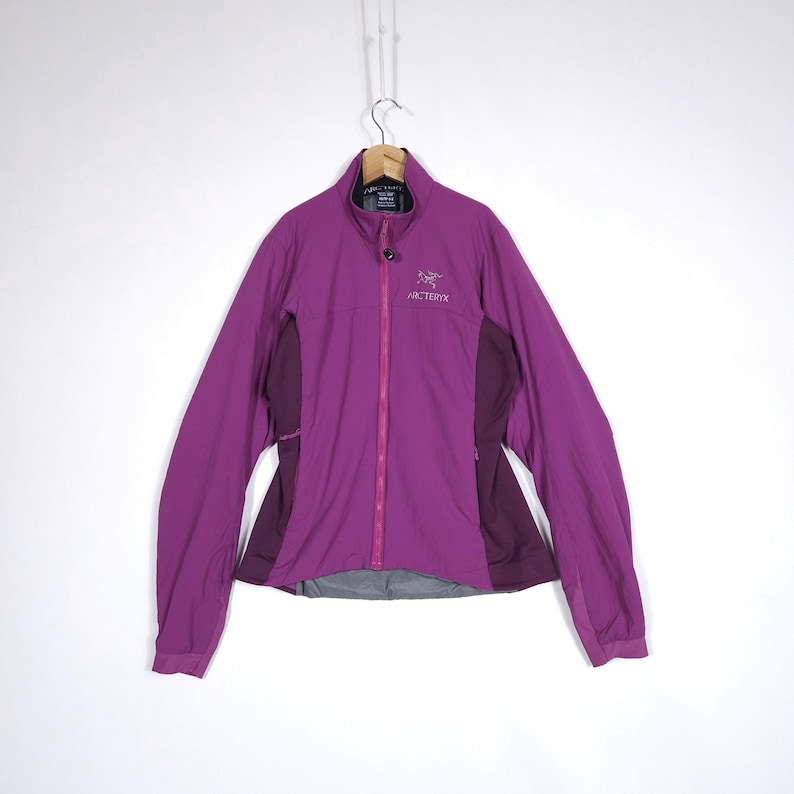 May include: A purple jacket with a full-length zipper and the brand name "Arc'teryx" on the chest. The jacket has a high collar and dark purple panels on the sides. It is hanging on a wooden hanger.