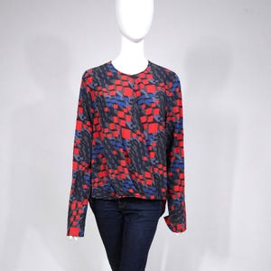 Puede incluir: Una blusa de manga larga con un patrón geométrico de cuadrados rojos, azules y grises sobre un fondo azul marino. La blusa tiene un cuello redondo y se muestra en un maniquí.