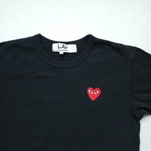 Peut inclure: T-shirt noir à col rond avec un écusson rouge en forme de cœur avec deux yeux blancs. Le t-shirt porte une petite étiquette blanche au niveau de l'encolure avec le texte "LAY des garcons".