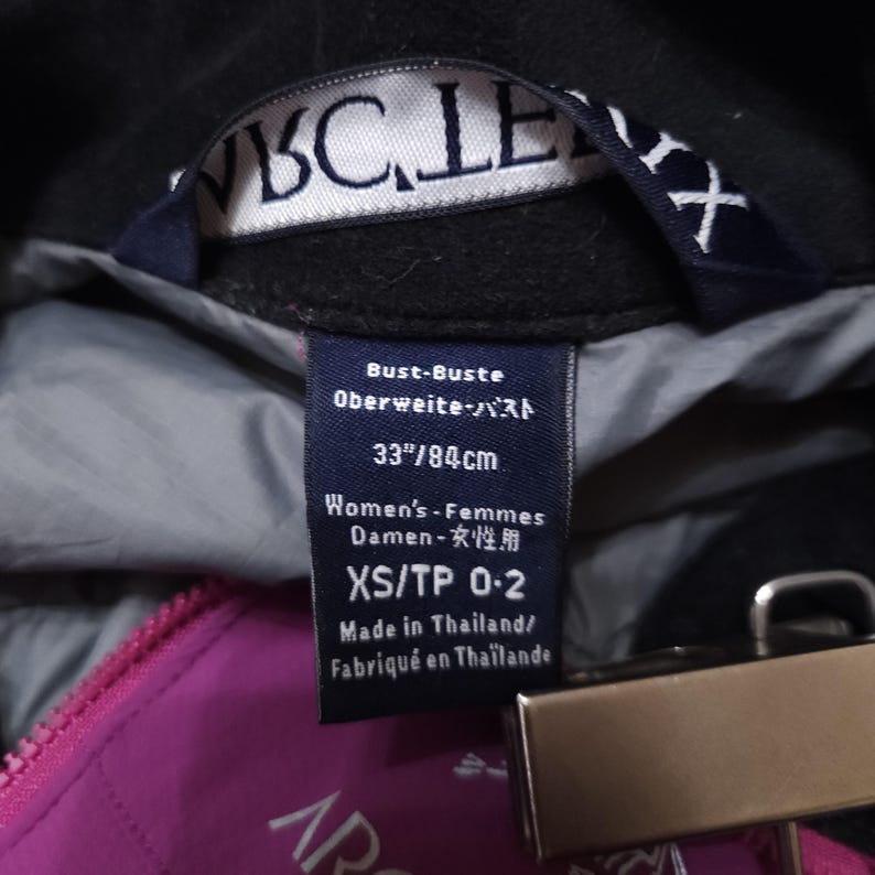 May include: Close-up of a black jacket's interior with a tag showing measurements of 33 inches or 84 cm. The tag also indicates the jacket is an XS/TP 0-2 size and made in Thailand. A pink zipper is visible.